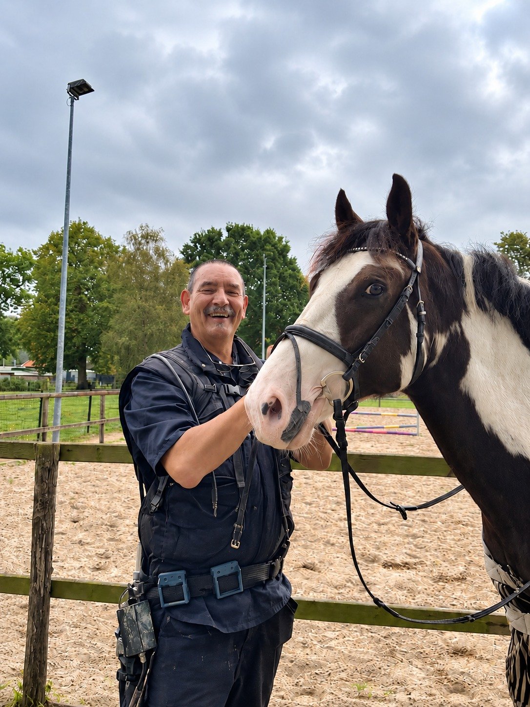 Marnix Brandes staat lachend naast een zwartbont paard met hoofdstel bij een buitenrijbaan.