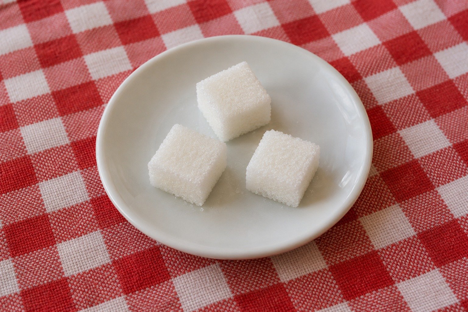 Drie witte suikerklontjes op een wit schoteltje op een rood-wit geruit tafelkleed
